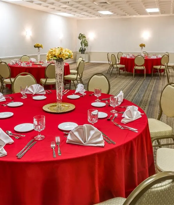 Sala de eventos con mesas redondas vestidas de rojo, decoradas con servilletas blancas y cubiertos.