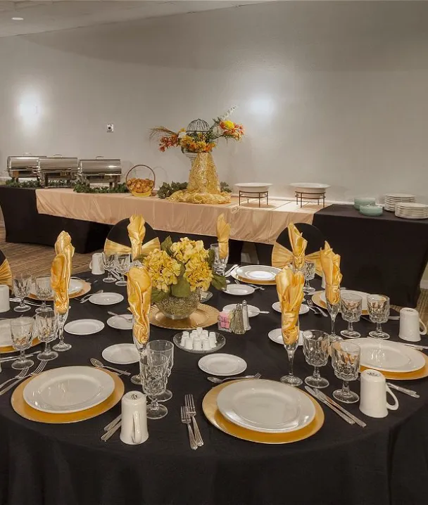 Mesa elegante con manteles negros y servilletas doradas, mesa de bufé al fondo.