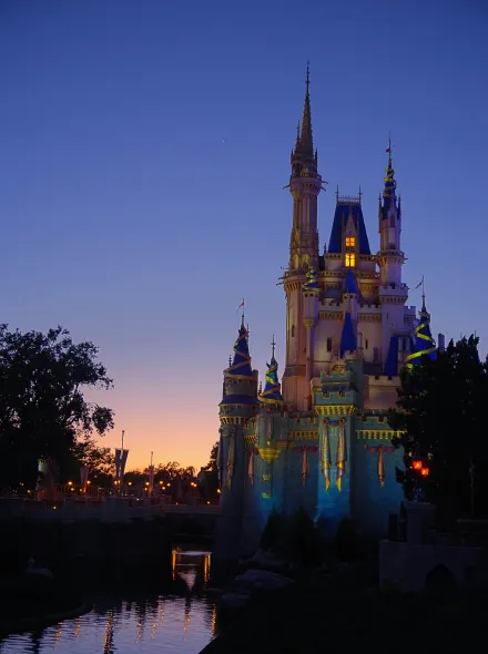 Castillo iluminado al atardecer con cielo azul, rodeado de &aacute;rboles y reflejado en el agua.