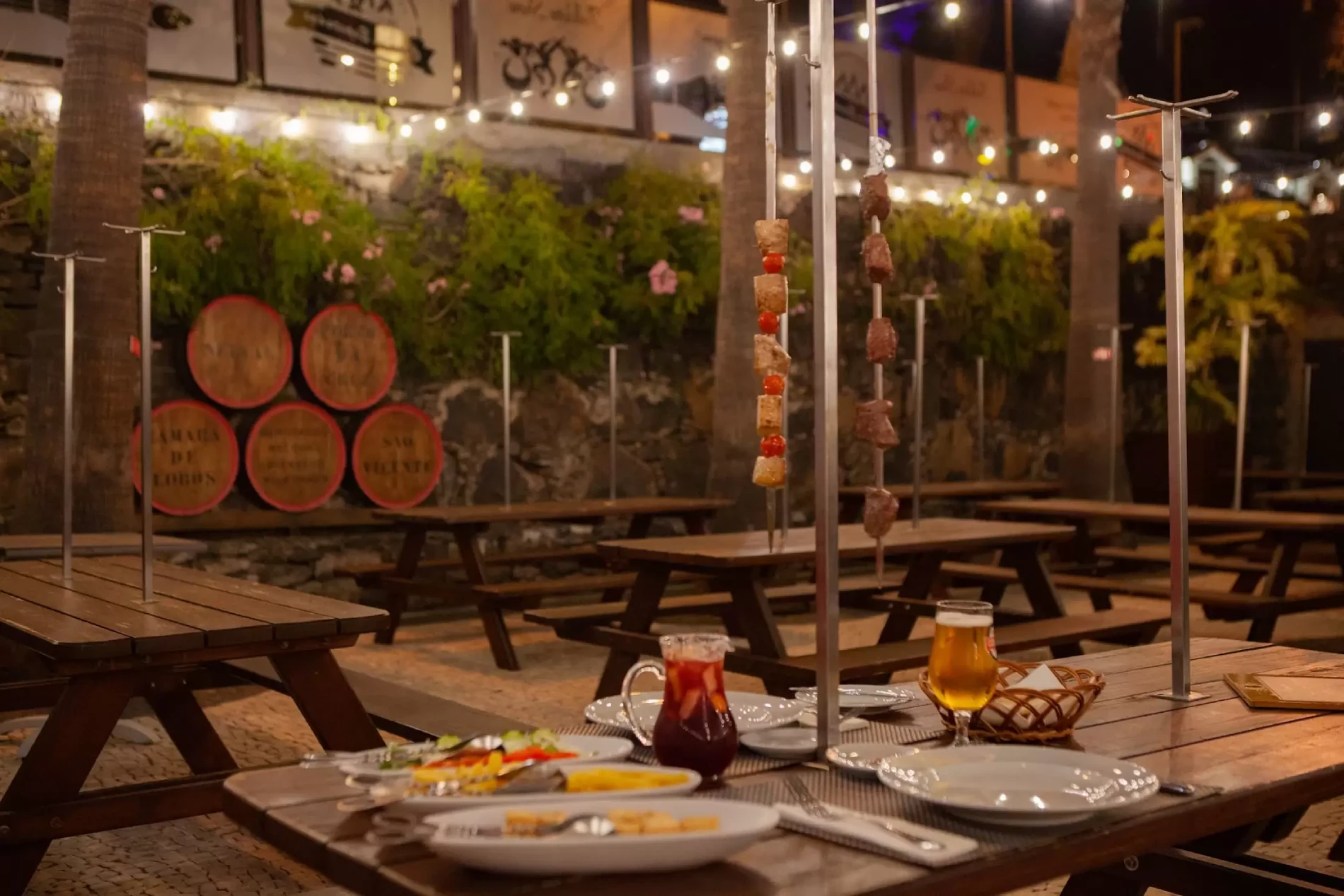 Outdoor dining area with skewers, drinks, and string lights.
