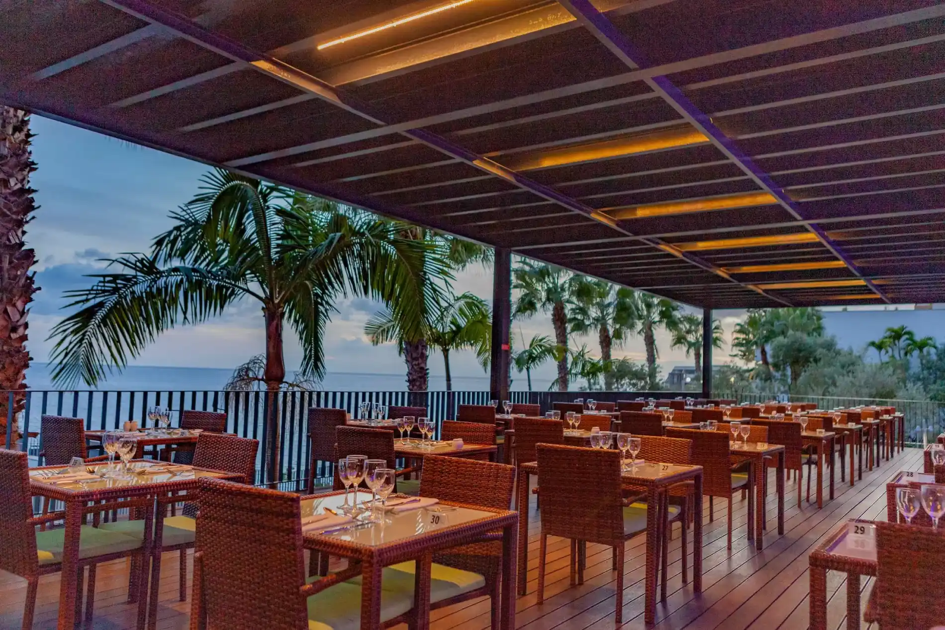 Outdoor restaurant with tables, chairs, palm trees, and ocean view at sunset.