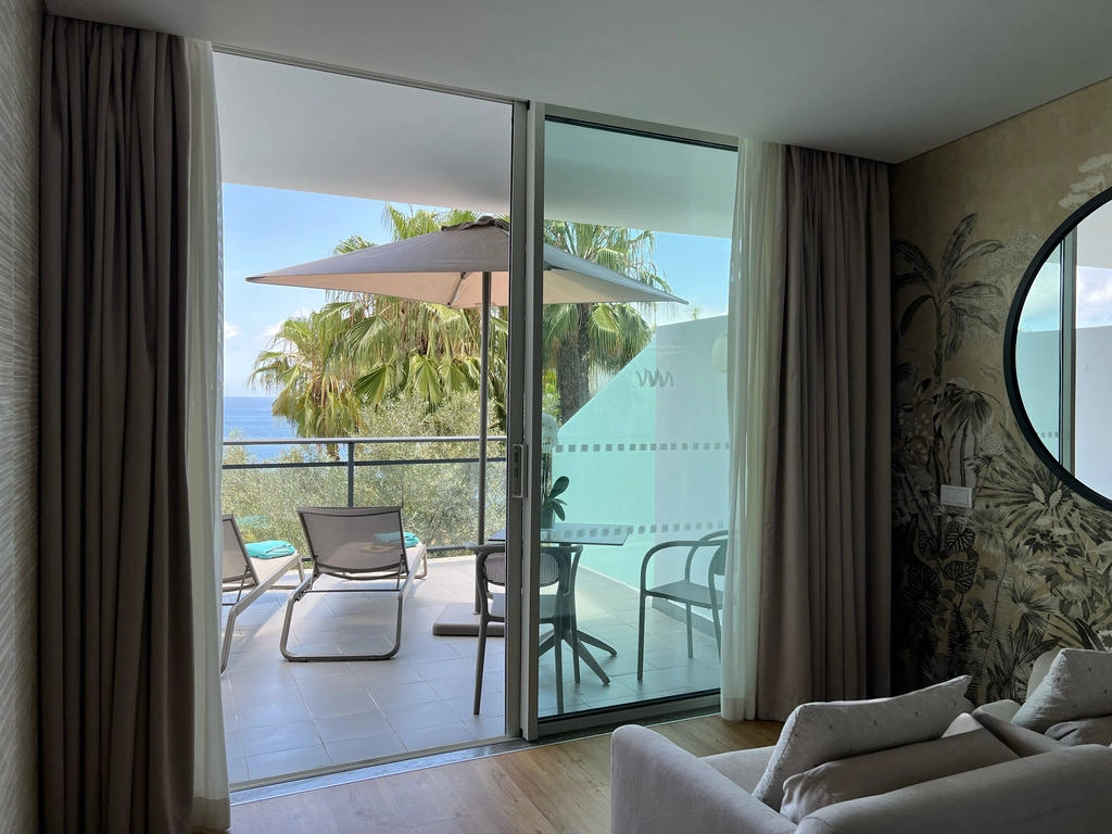 Room with balcony, chairs, umbrella, and a sea view through glass doors.