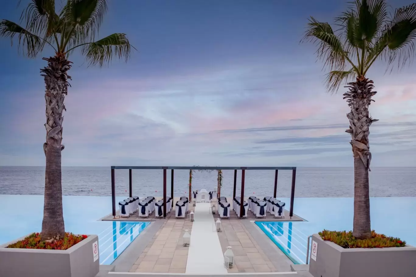 Oceanfront wedding setup with palm trees and infinity pool at sunset.