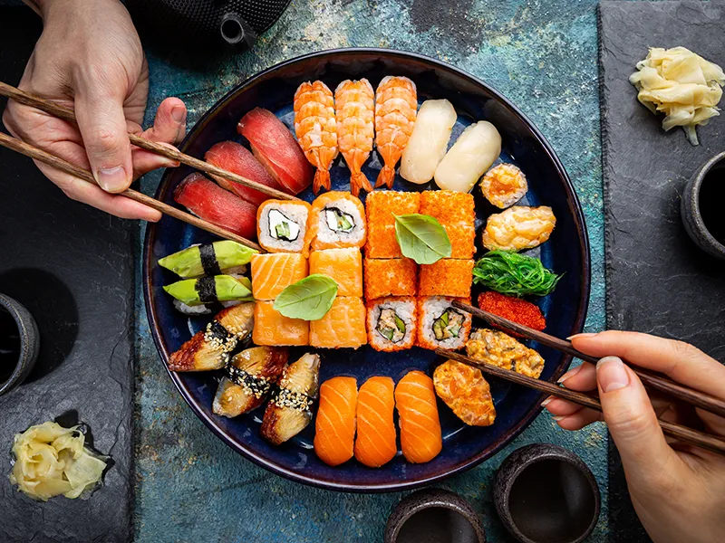 Hands holding chopsticks above a plate of assorted sushi.