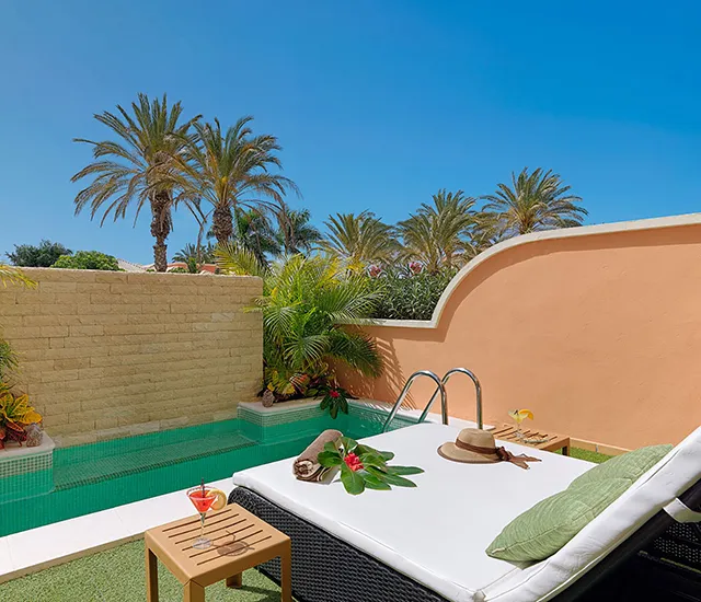 Poolside lounge chair with hat, drink, and palm trees under a clear blue sky.