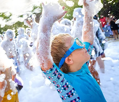 Child in goggles enjoying a foam party, arms raised amidst bubbles.