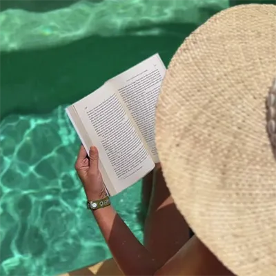 Person reading by a pool, wearing a hat.