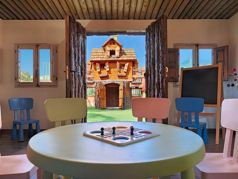 Children's playroom with a round table and chairs, view of a wooden playhouse outside.
