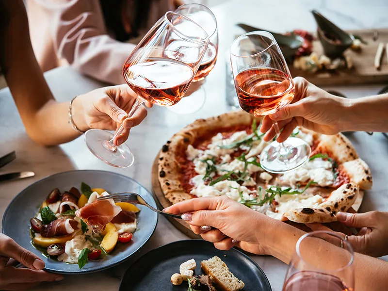 Hands toasting with wine over pizza and appetizers on a table.
