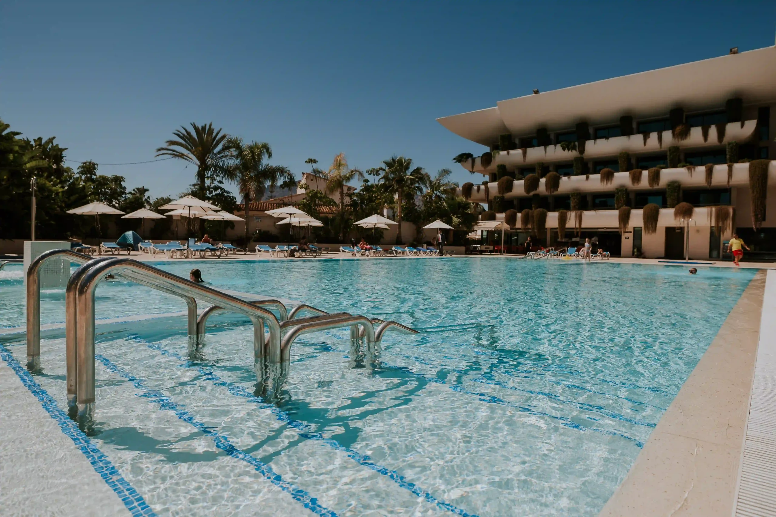 Piscina de hotel con escaleras met&aacute;licas y sombrillas blancas al fondo.