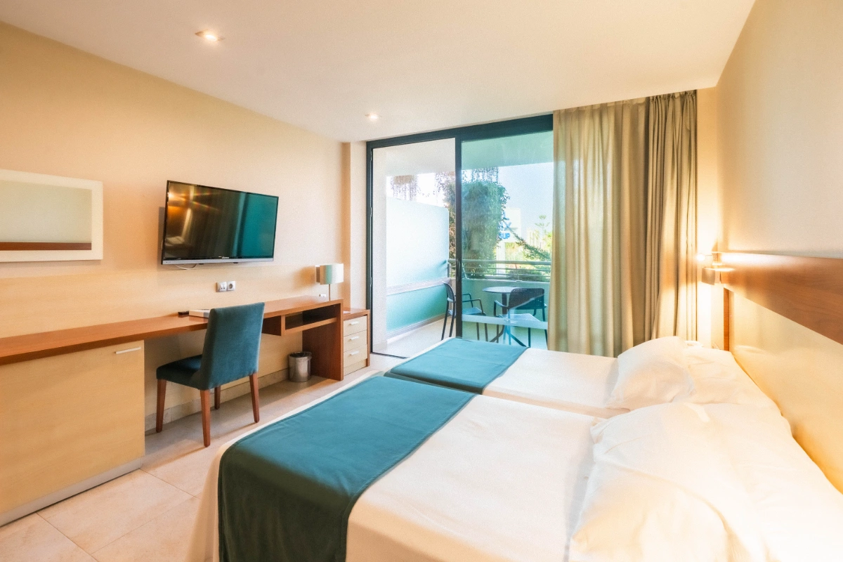 Habitaci&oacute;n de hotel con dos camas, televisor y balc&oacute;n con vista exterior.