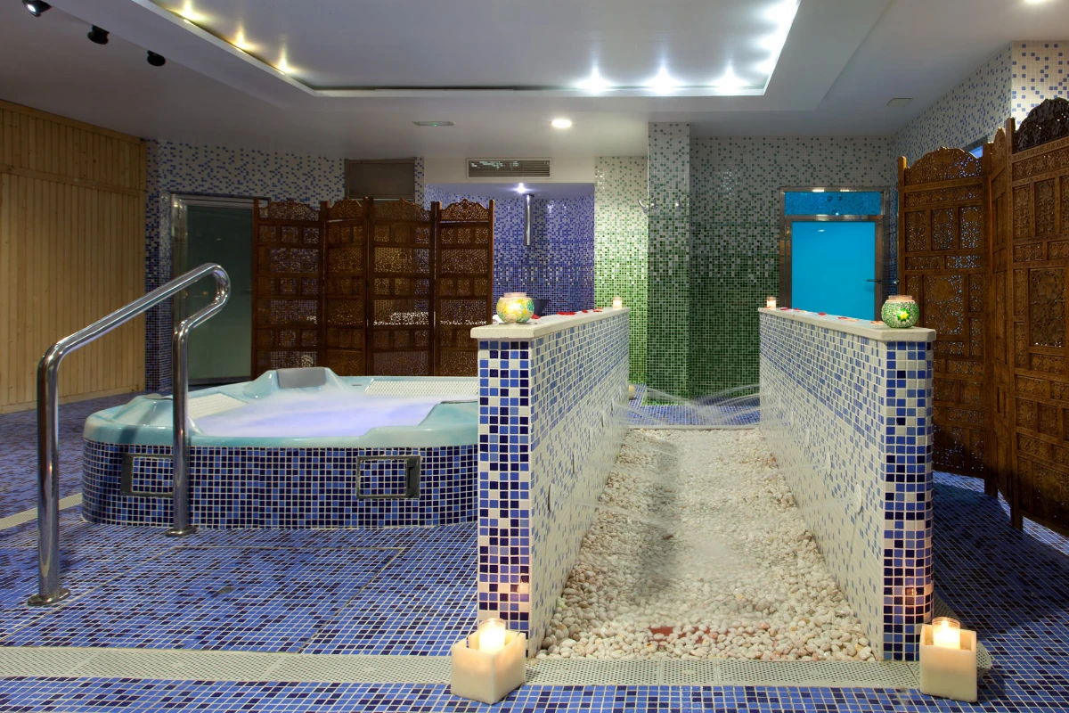 Sala de spa con jacuzzi, velas y paredes decoradas con mosaicos azules y verdes.