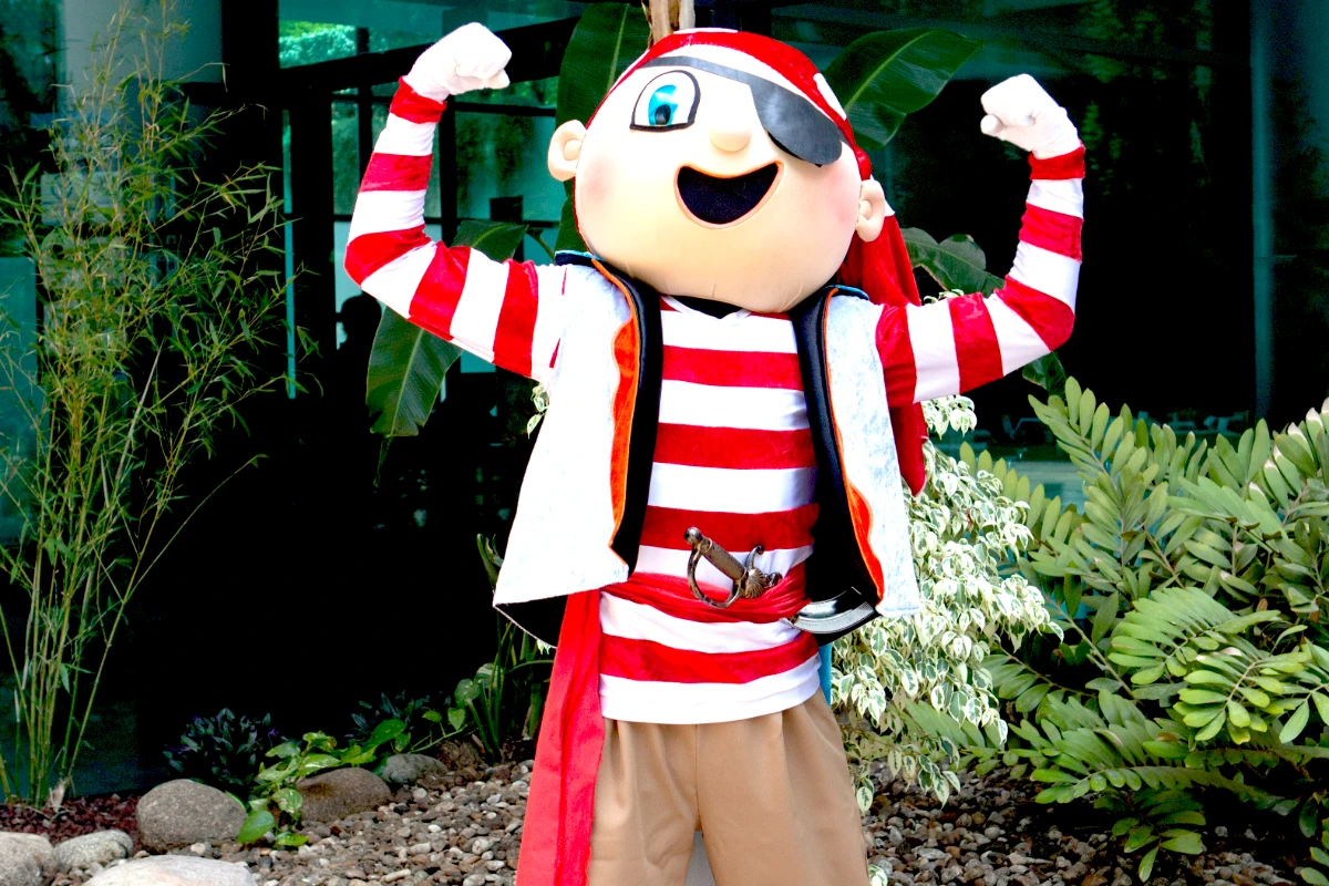 Personaje disfrazado de pirata, con los brazos en alto, rodeado de plantas.