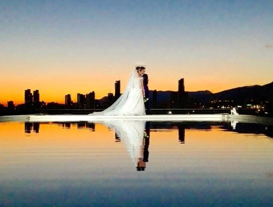 Novios de pie al atardecer junto a un estanque, reflejados en el agua.