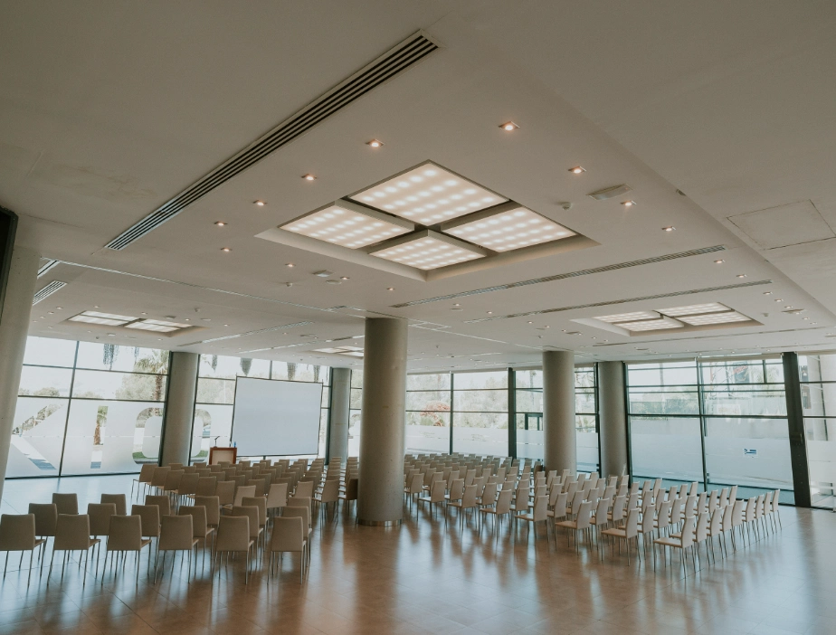 Sala de conferencias vac&iacute;a con sillas y pantallas, techo con luces modernas.
