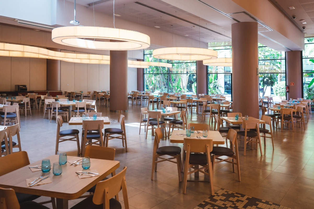 Comedor amplio con mesas y sillas de madera, luces circulares y ventanas grandes.