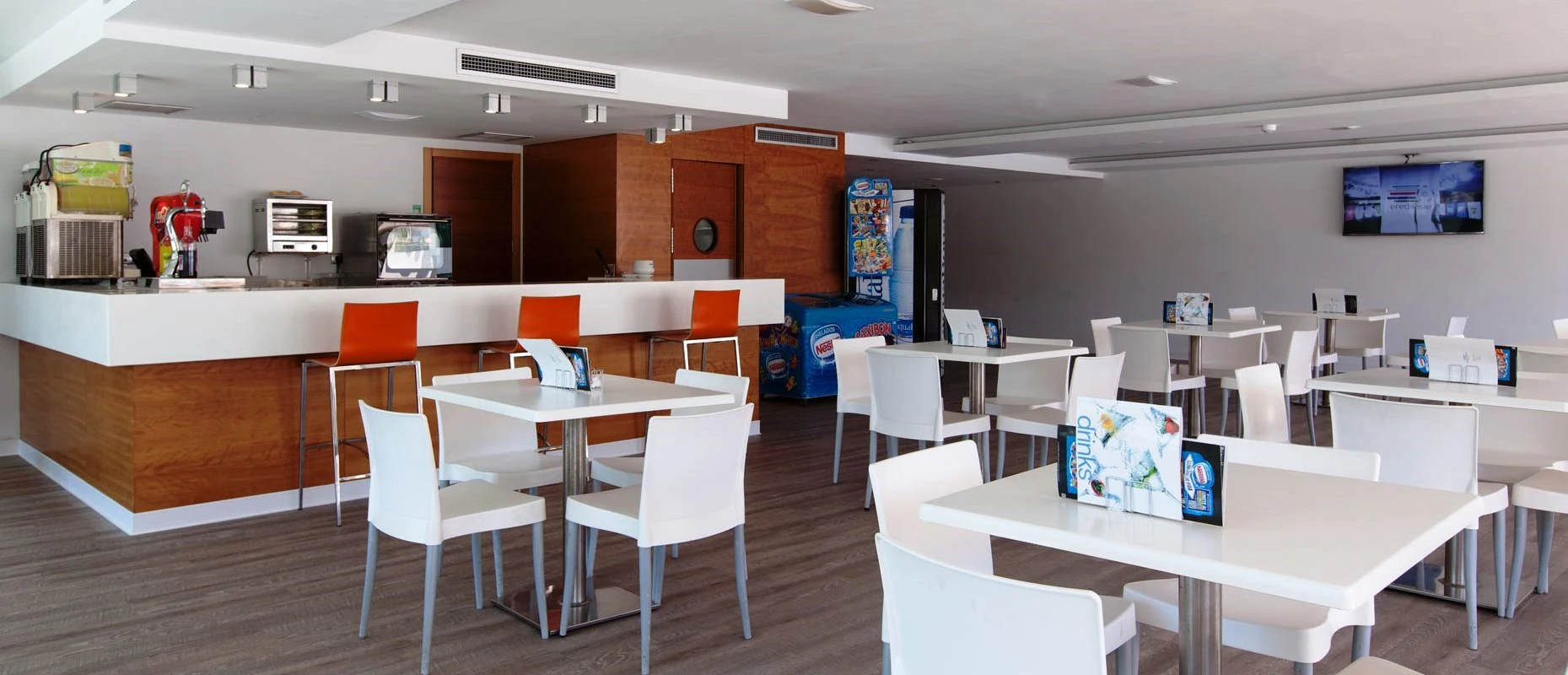 Cafeter&iacute;a moderna con mesas blancas y sillas naranjas frente a un mostrador.