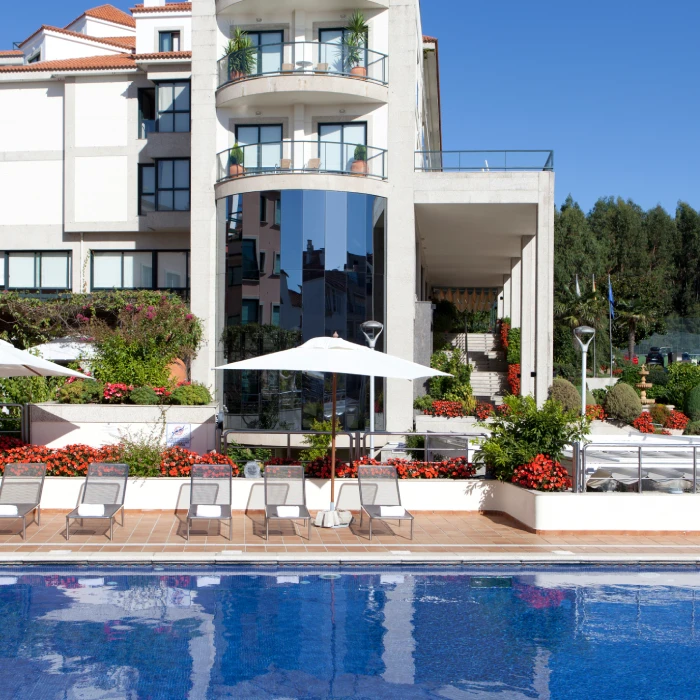 Edificio moderno con piscina, sillas y jardines florales bajo un cielo azul.