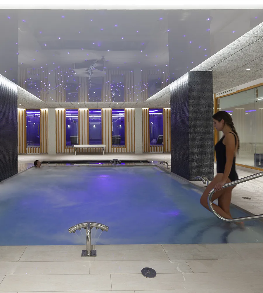 Piscina interior iluminada, con personas relaj&aacute;ndose en el agua.