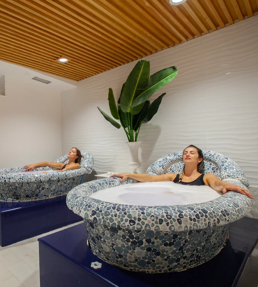 Mujeres se relajan en ba&ntilde;eras de piedra en un spa, con una planta al fondo.