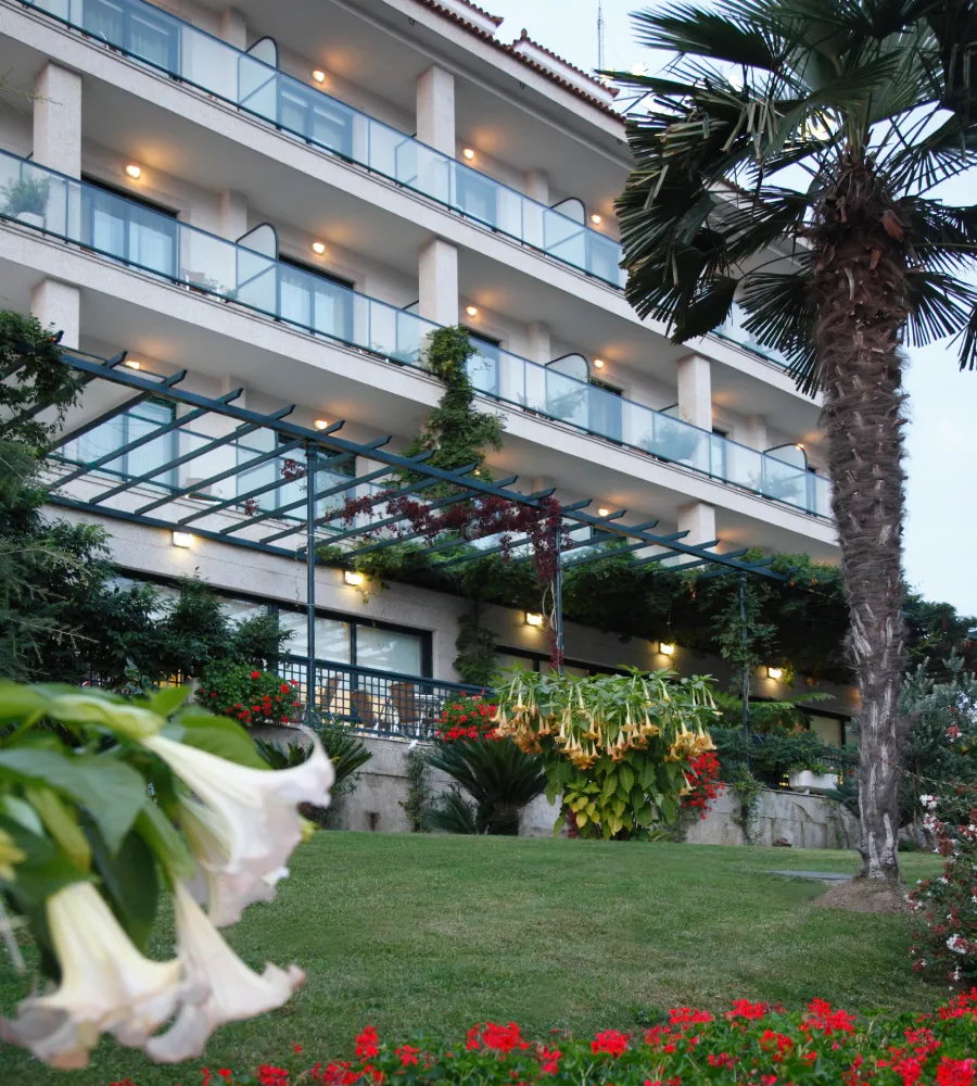 Hotel con balcones, jard&iacute;n florido y palmeras al frente.