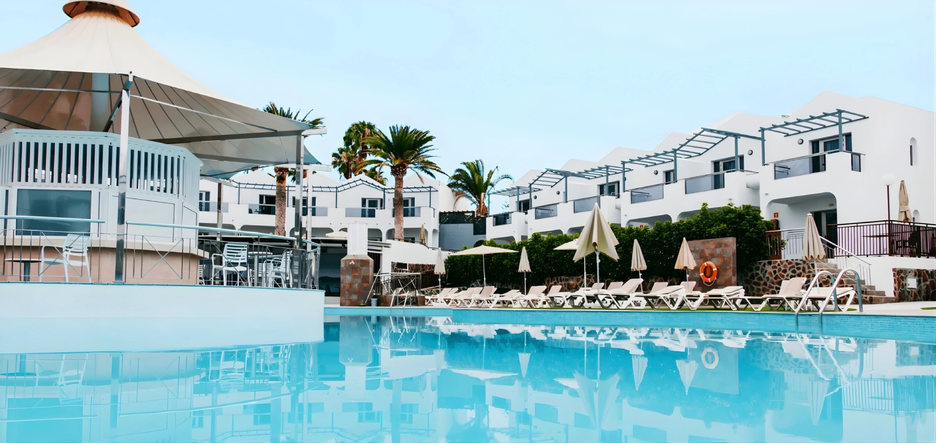 Piscina con tumbonas, sombrillas y edificio blanco al fondo.