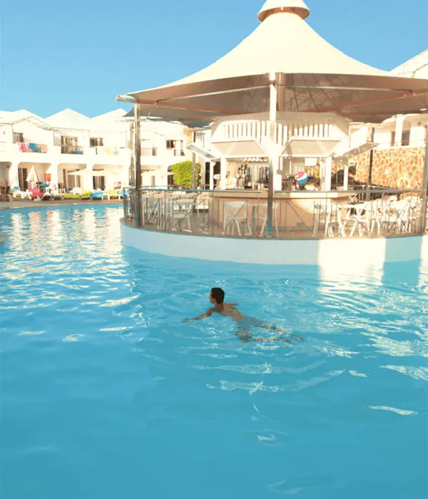 Hombre nadando en piscina al aire libre frente a bar con techo blanco.