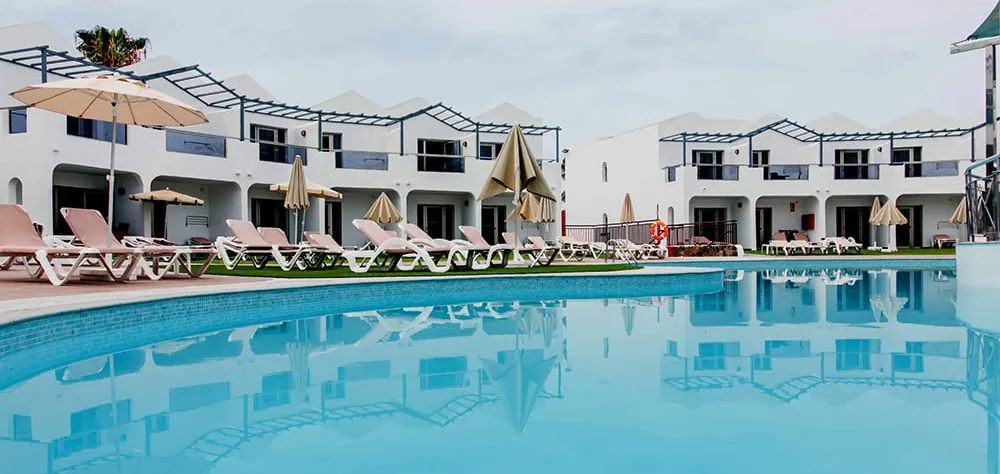 Piscina frente a un hotel blanco con tumbonas y sombrillas.