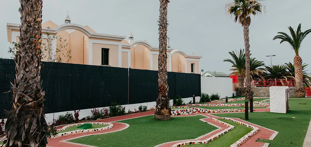 Minigolf rodeado de palmeras y una cerca, con edificios al fondo.