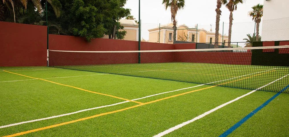 Cancha de tenis en c&eacute;sped artificial con casas y palmeras alrededor.