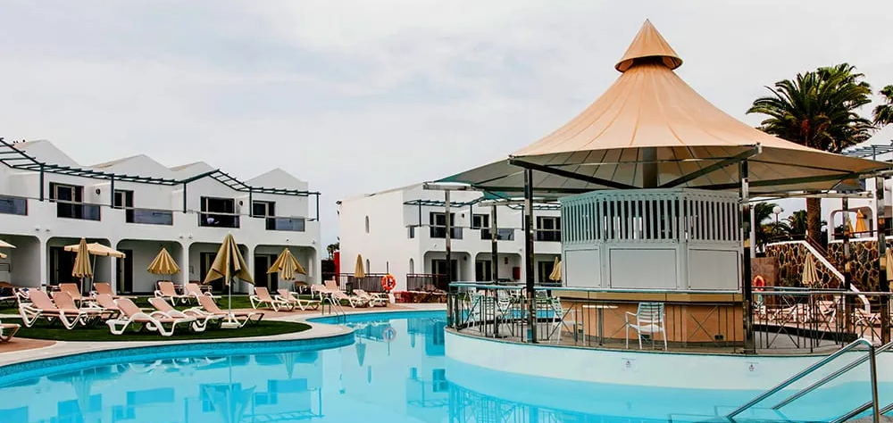 Piscina y sombrillas junto a edificios blancos y un kiosco cubierto.