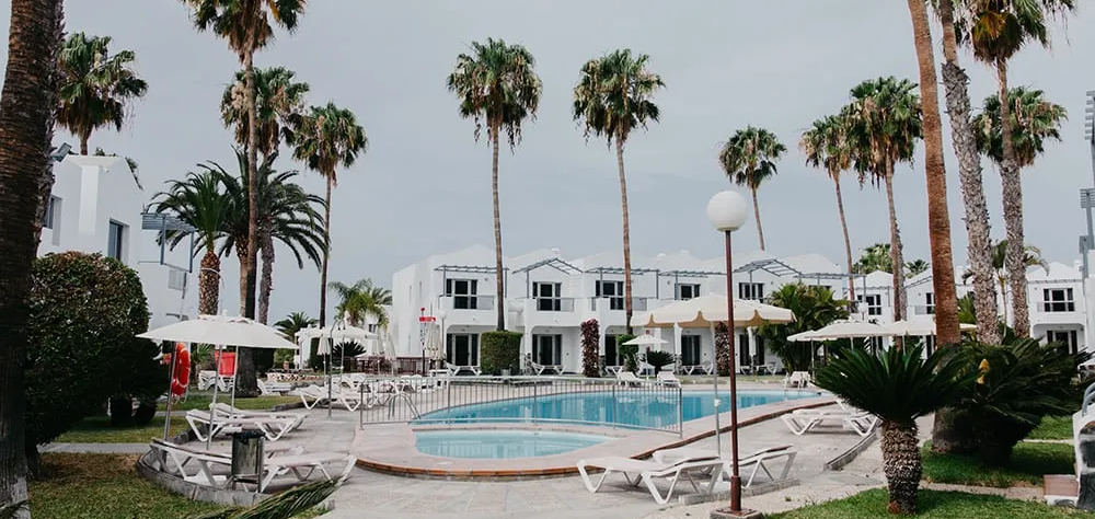 &Aacute;rea de piscina rodeada de casas blancas y palmeras bajo un cielo nublado.