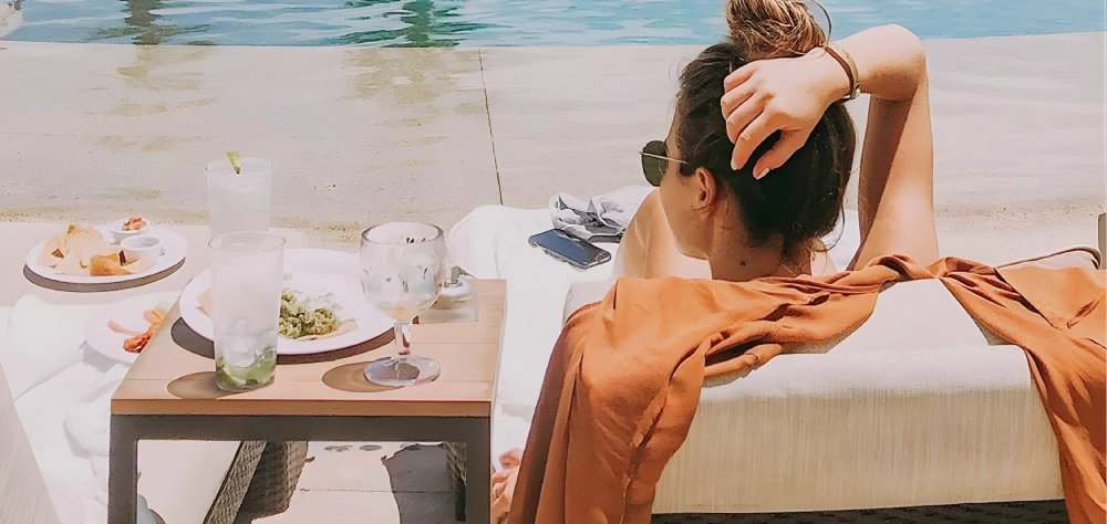 Mujer relaj&aacute;ndose junto a una piscina, con mesa de bebidas y aperitivos.