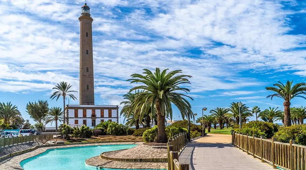 Faro junto a palmeras y un camino; cielo azul con nubes.