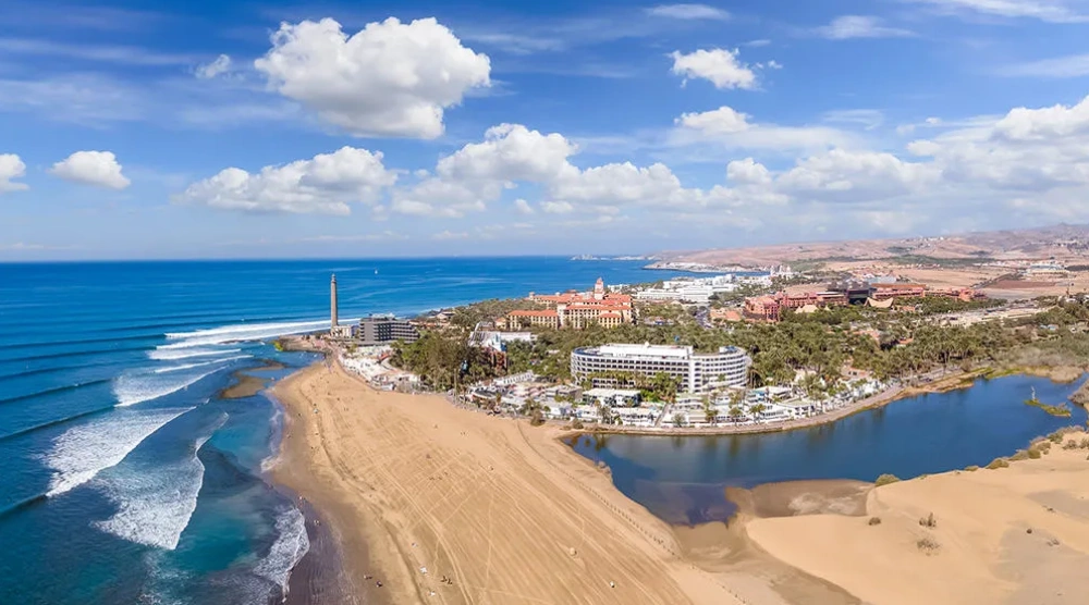 Vista a&eacute;rea de una playa con un faro y edificios. Cielo azul con nubes.