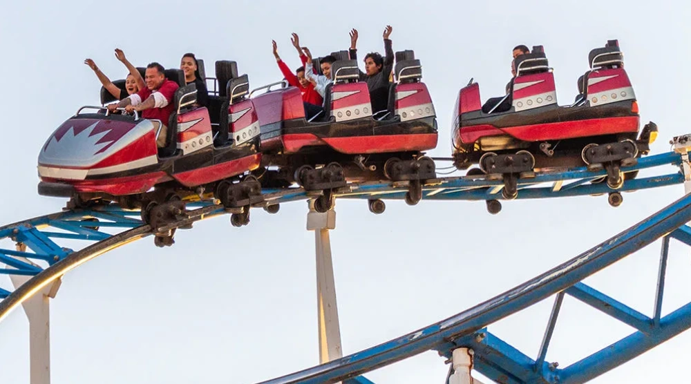 Personas disfrutando una monta&ntilde;a rusa con los brazos levantados al atardecer.