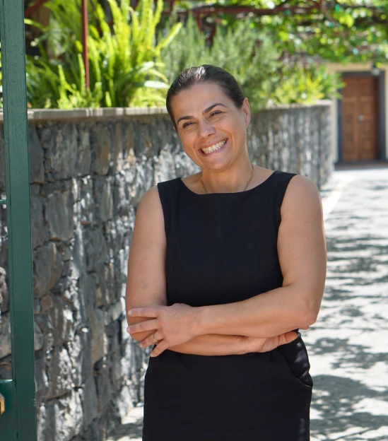 Smiling woman in a black dress stands by a stone wall outdoors.