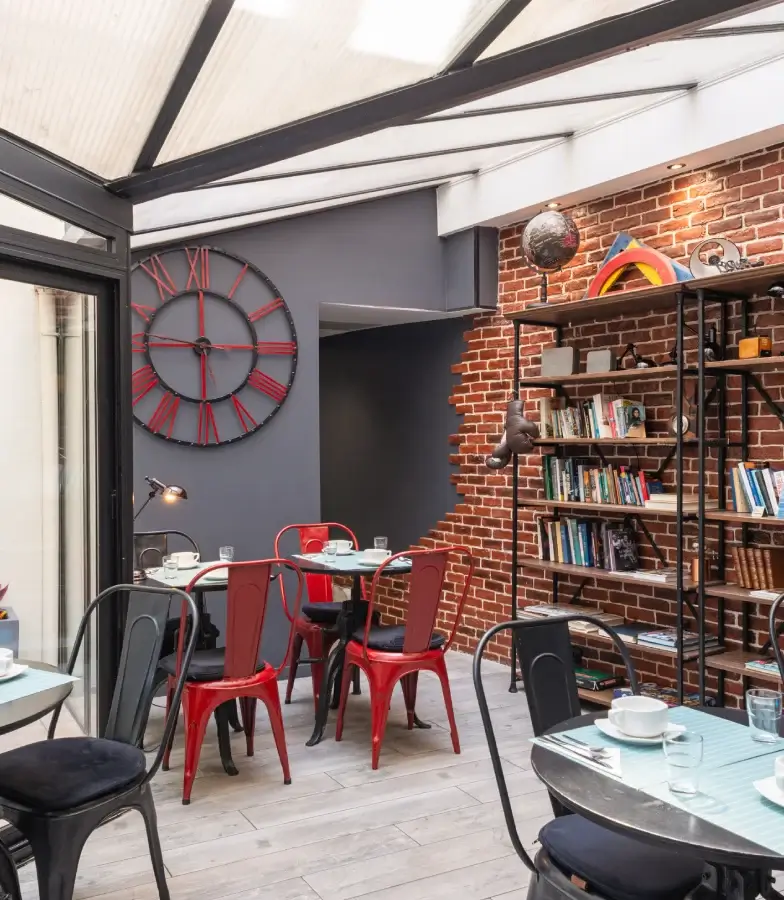 Patio int&eacute;rieur avec chaises rouges, tables, horloge murale et briques apparentes.