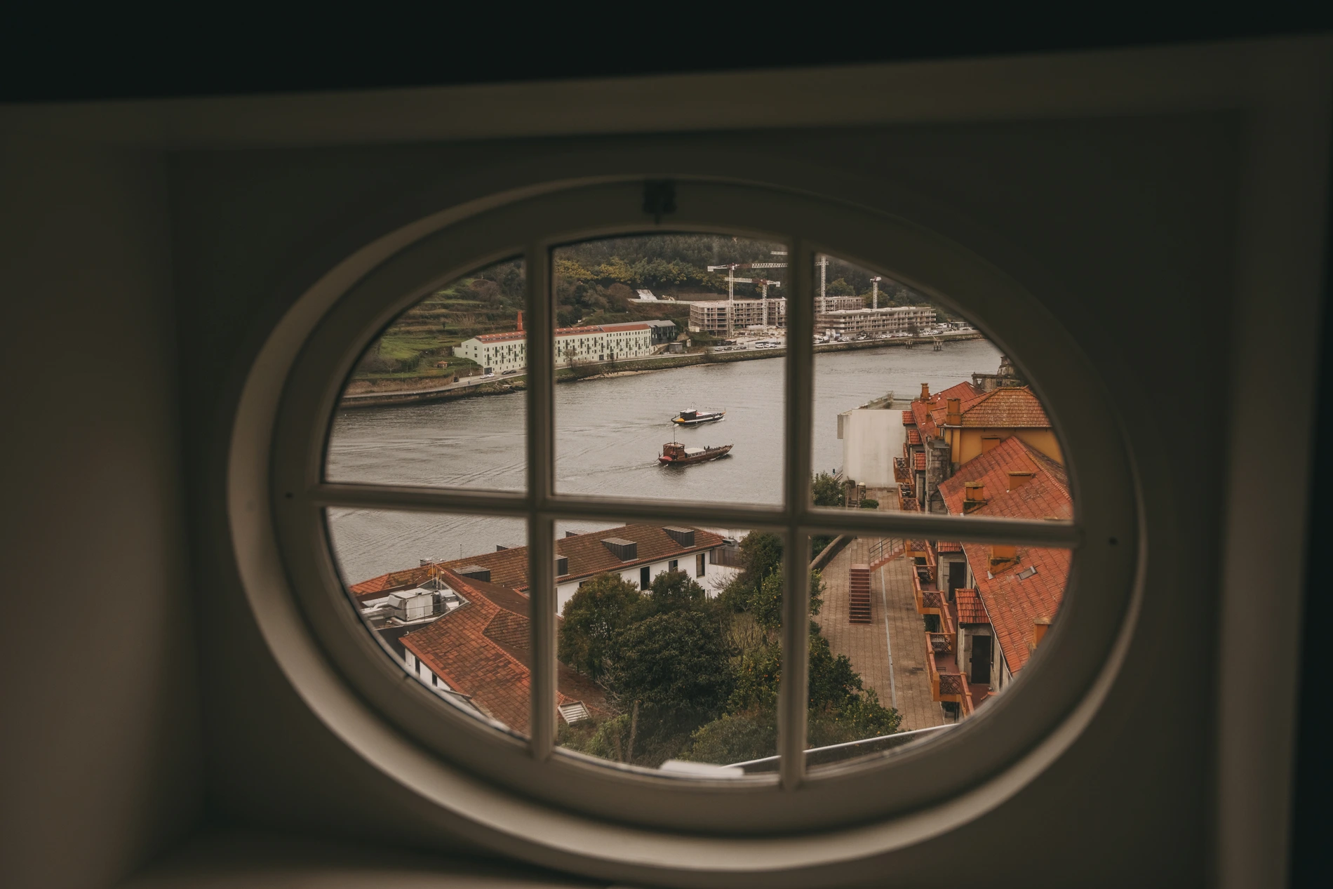 Oval window view of a river with boats and orange-roofed buildings.