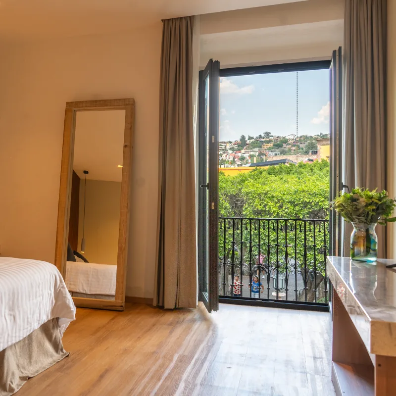 Habitaci&oacute;n con cama, espejo, balc&oacute;n abierto y vista a &aacute;rboles y casas.