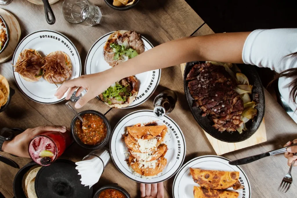 Mesa llena de platos variados en un restaurante, con manos sirviendo comida.