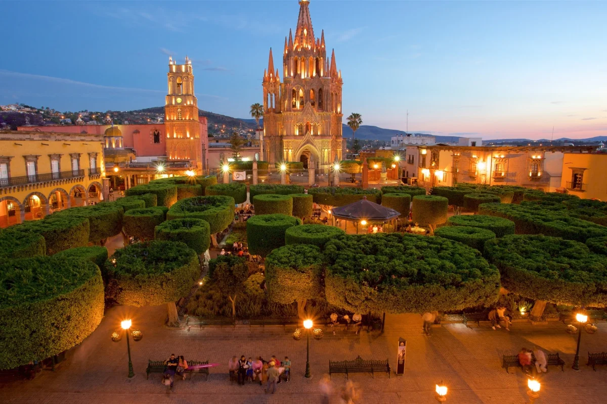Plaza con jardines y una iglesia g&oacute;tica iluminada al atardecer.