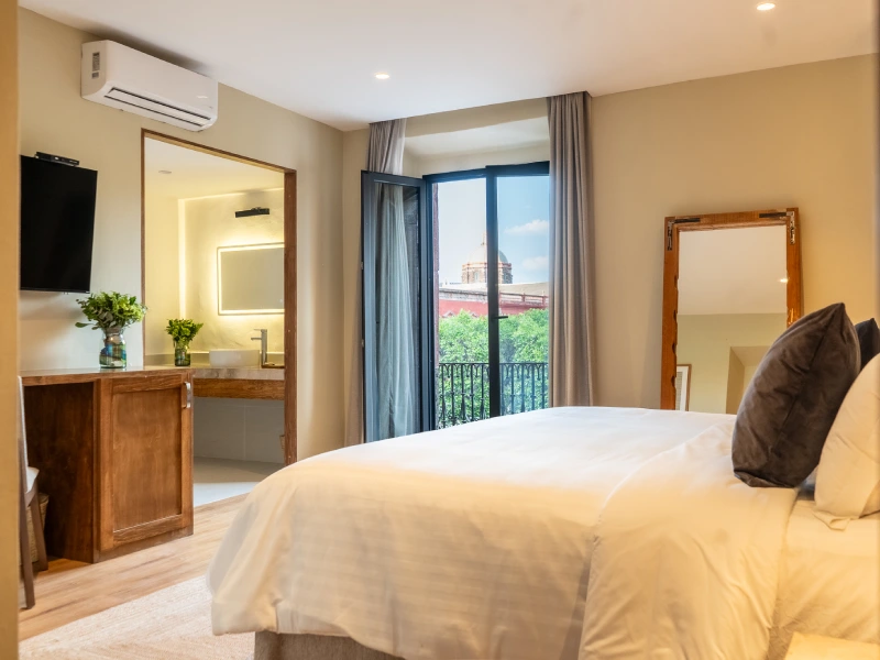 Habitaci&oacute;n moderna con cama, espejo, balc&oacute;n y ba&ntilde;o iluminado.