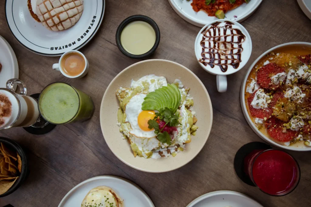 Desayuno variado con chilaquiles, bebidas y postres en una mesa de madera.