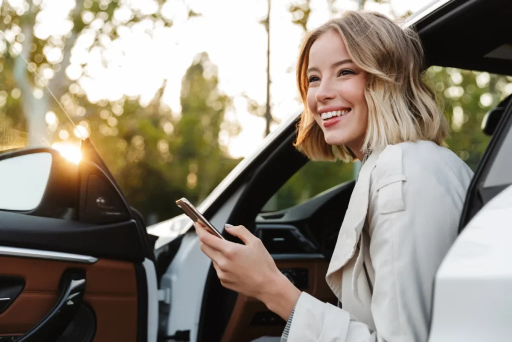 Mujer sonriente con m&oacute;vil junto a un coche al atardecer.