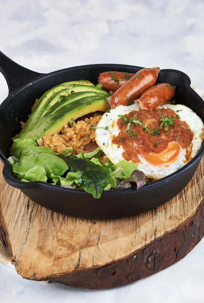 Sart&eacute;n con huevo, salchichas, aguacate y ensalada sobre tabla de madera.