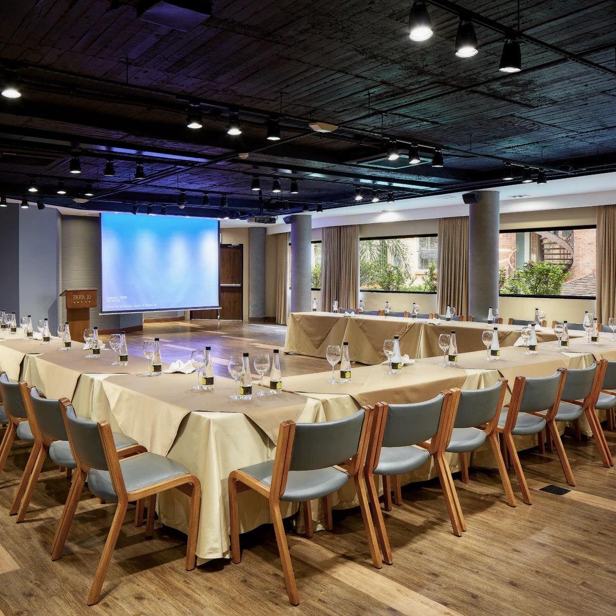 Sala de conferencias con mesas en U y pantalla de proyecci&oacute;n encendida.