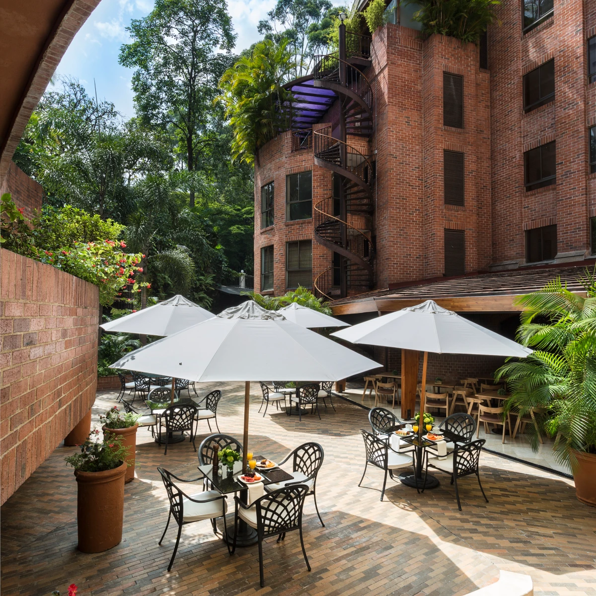 Patio con mesas y sombrillas rodeado de vegetaci&oacute;n y edificio de ladrillo.