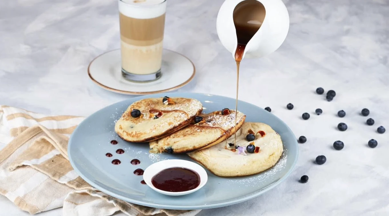 Plato con panqueques y ar&aacute;ndanos, acompa&ntilde;ado de caf&eacute; y jarabe por encima.