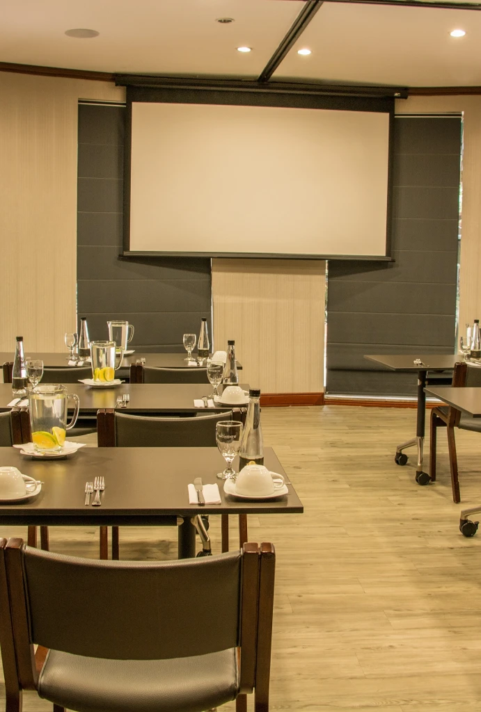 Sala de reuniones con mesas, sillas y pantalla de proyecci&oacute;n.